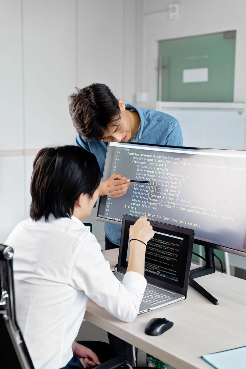 Two engineers reviewing application code together across monitors in a bright office