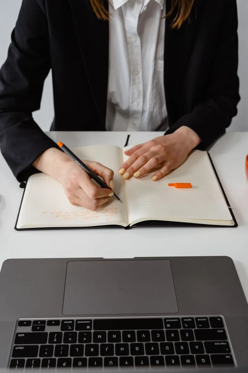 A businesswoman taking notes at a laptop and notebook on a bright office desk