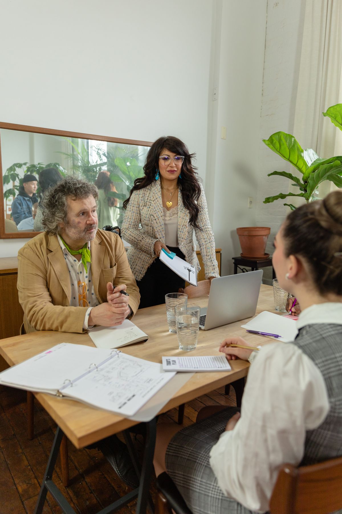 Three colleagues talking around a wooden table with a laptop and documents in a bright office, planning a financial services security project