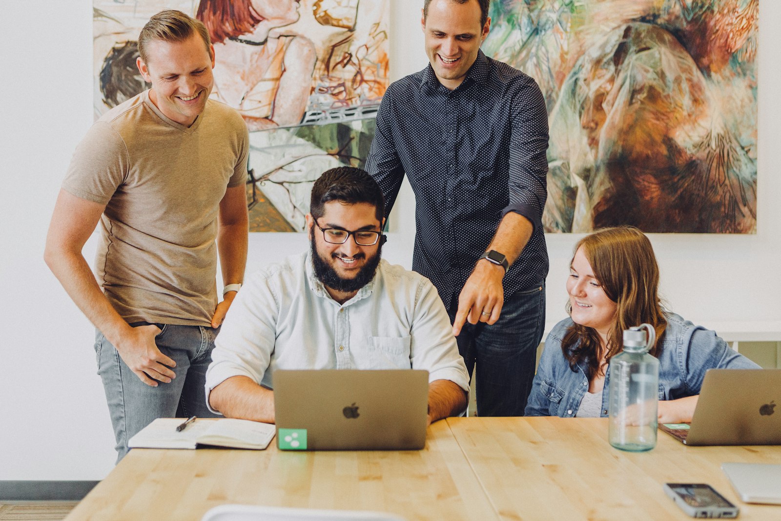 Four smiling colleagues gathered around a laptop at a wooden table with artwork on the wall