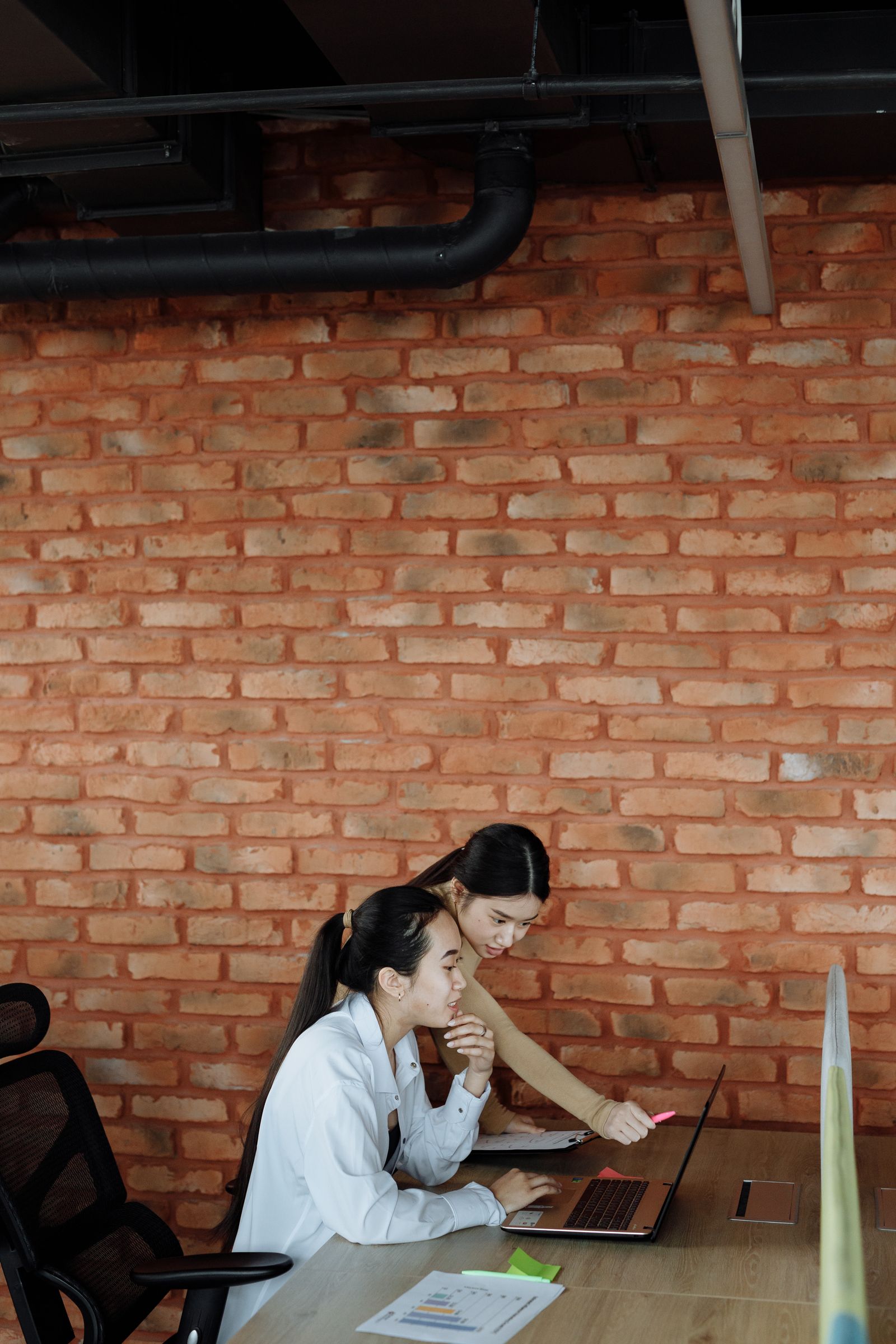 Two women reviewing a laptop at a desk against an exposed brick wall