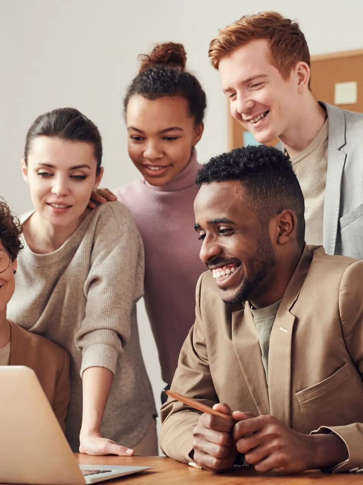 A diverse group of colleagues smiling around a laptop at a wooden table