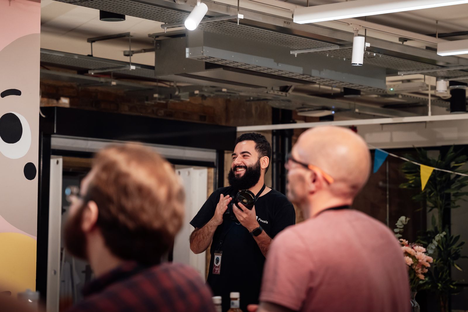 A smiling bearded man holding a camera in a modern office with colleagues in the foreground