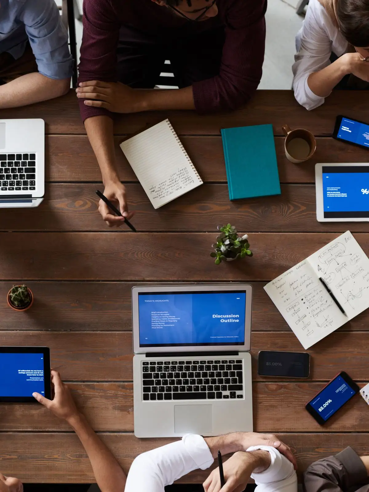 Overhead view of a wooden table with laptops, tablets and notebooks surrounded by colleagues' hands