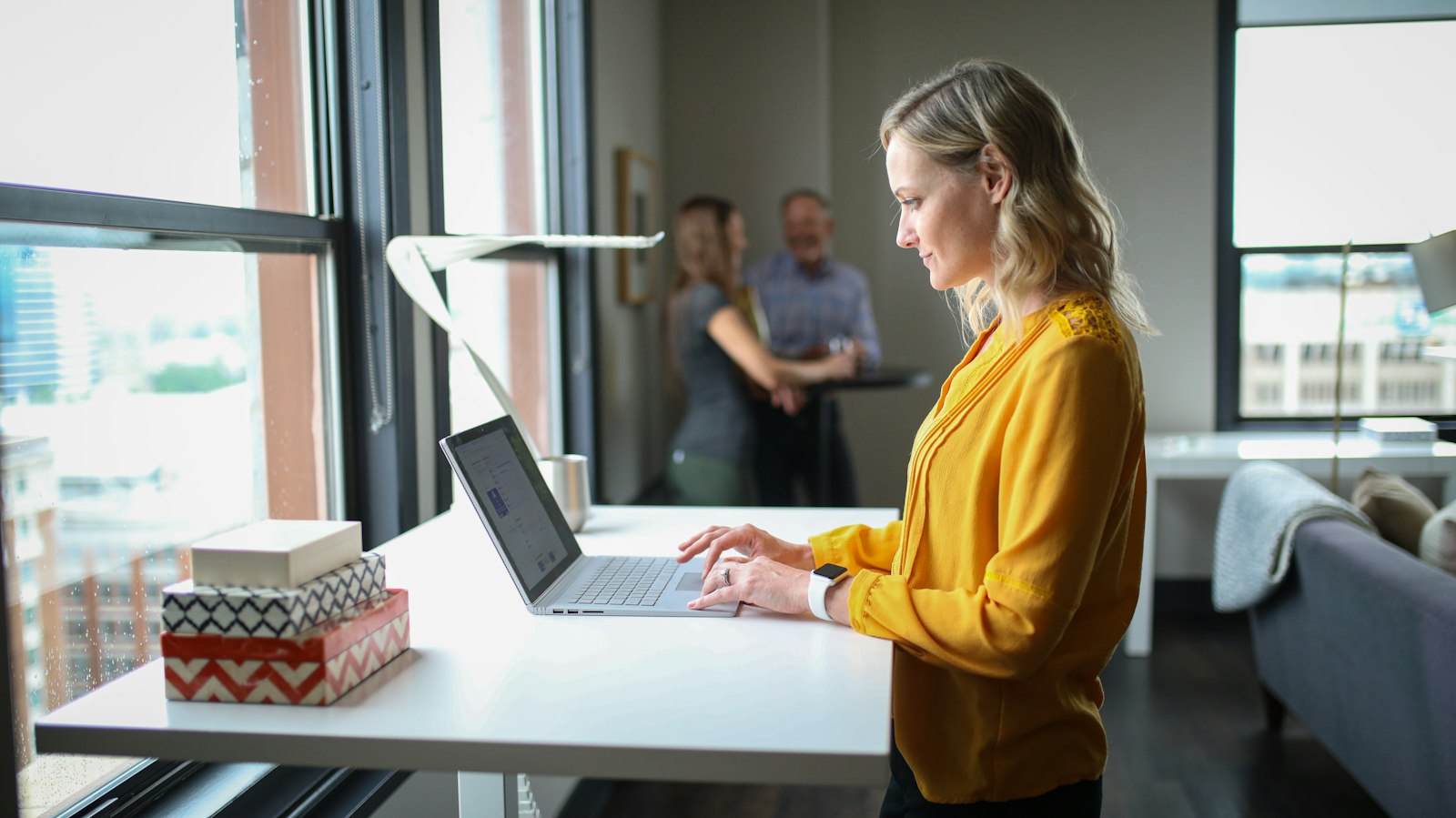 A woman in a yellow blouse typing at a standing desk beside a large window
