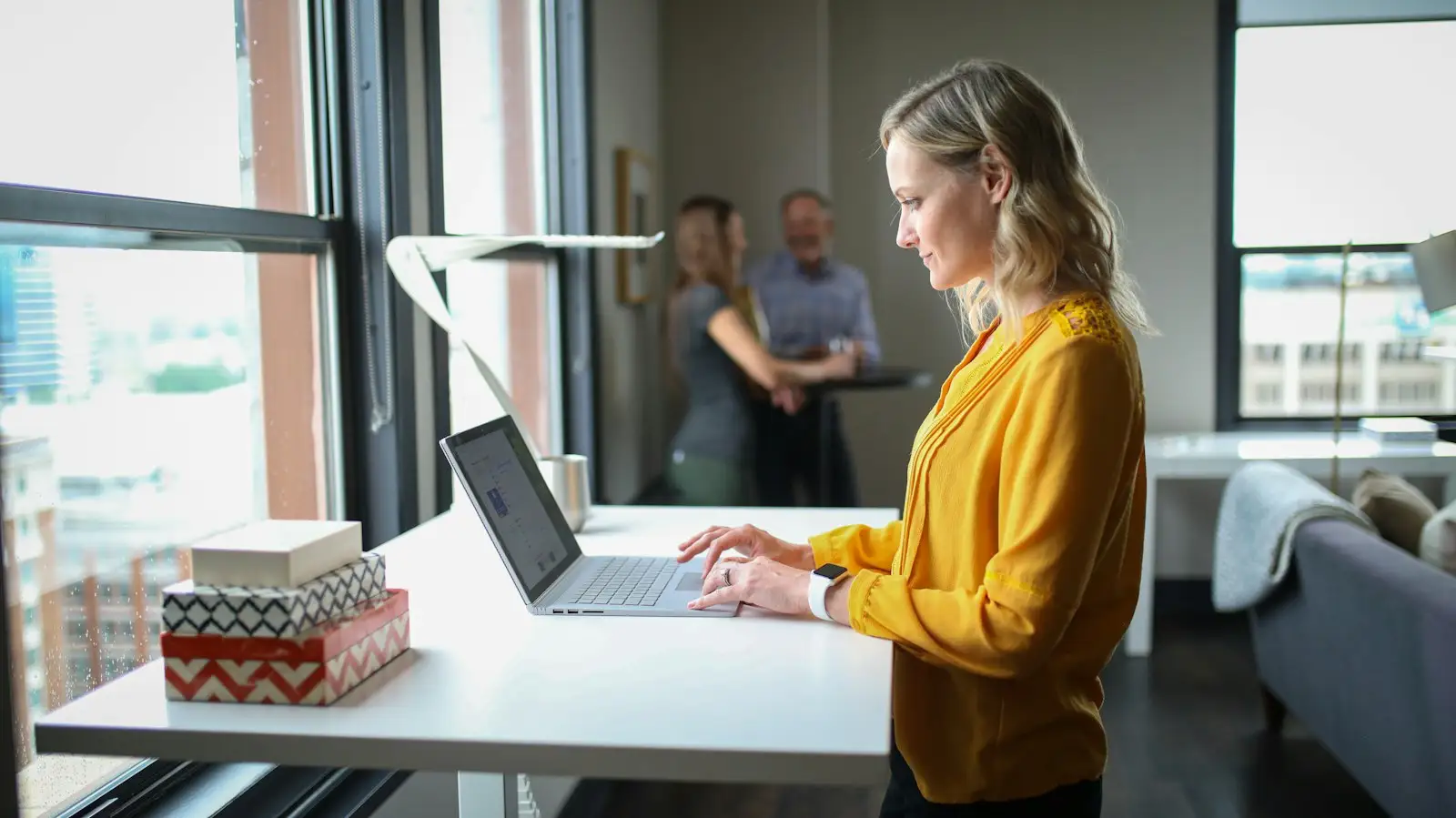 A woman in a yellow blouse typing at a standing desk beside a large window