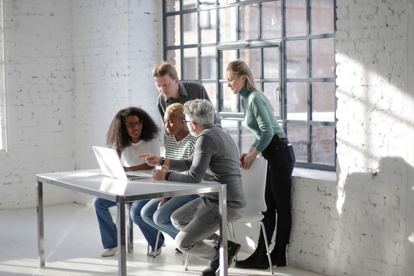 A diverse group of five colleagues gathered around a laptop at a glass table in a bright white brick office